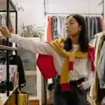Mains fouillant parmi des vêtements vintage et objets de collection sur les étagères d'une friperie.