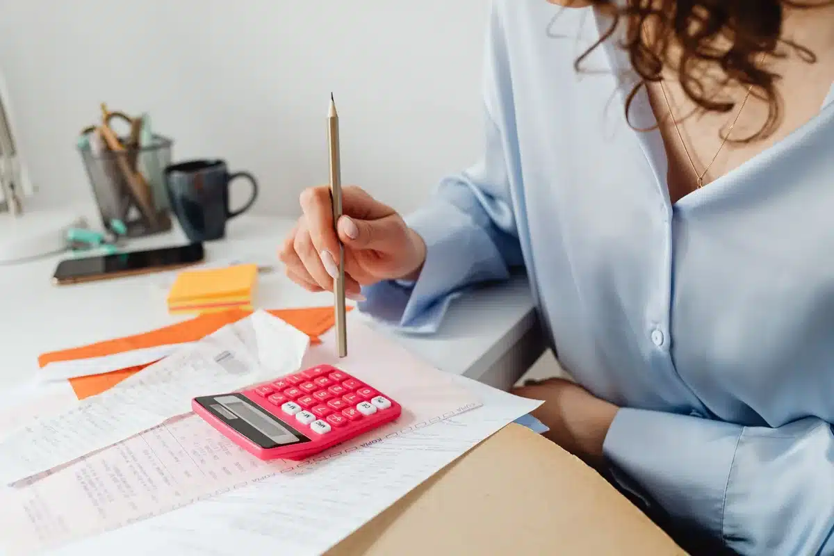 Personne rédigeant un budget sur un carnet avec calculatrice sur une table ensoleillée